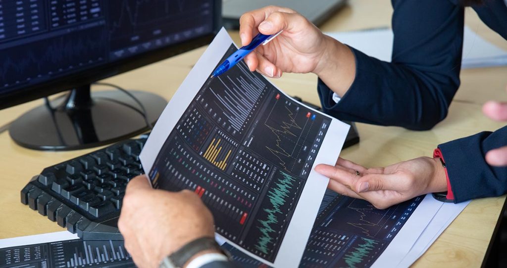 Két ember nyomtatott árfolyamgrafikonokat elemez közösen az irodai monitorok előtt