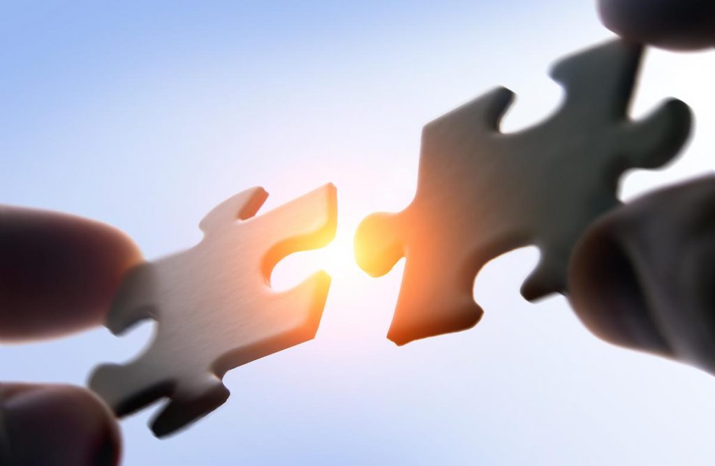 Két kéz illeszt össze két puzzle-darabot, mögöttük napfény